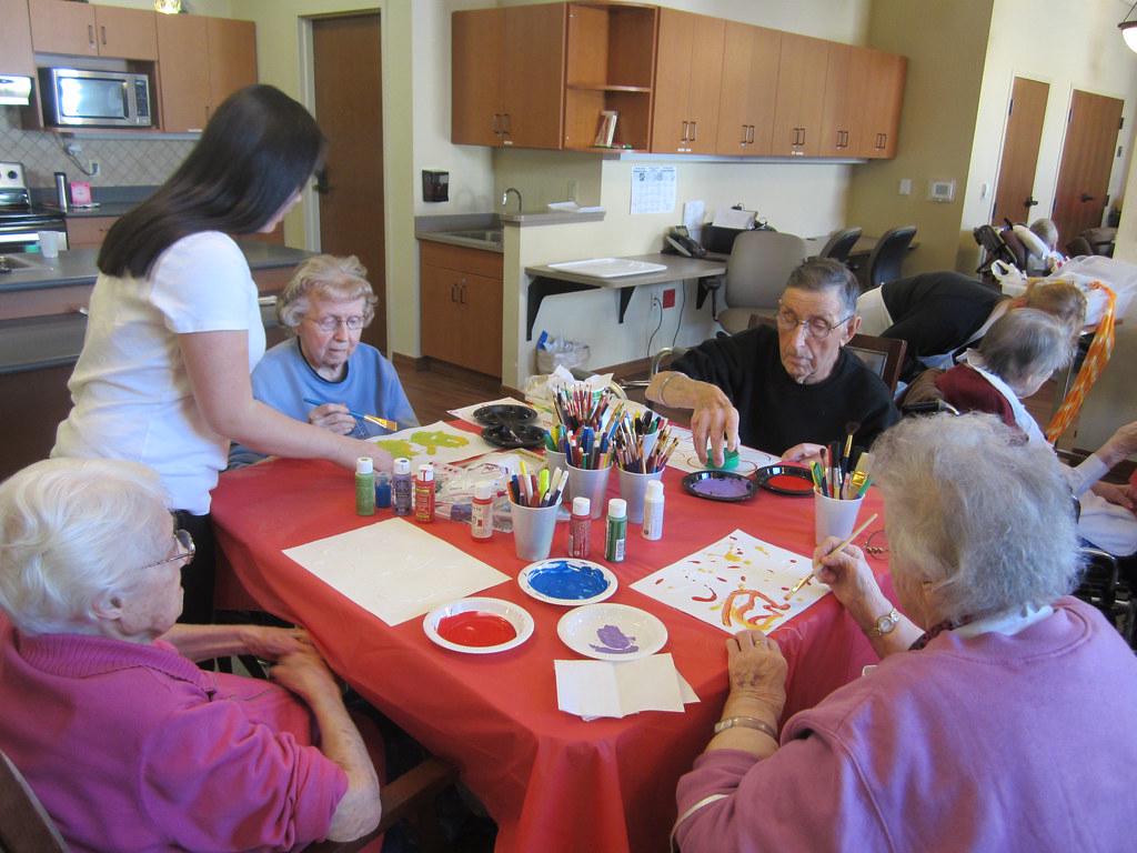 A group of seniors enjoying an art therapy session, painting on canvases and socializing with each other. The image conveys a sense of engagement, creativity, and social connection.